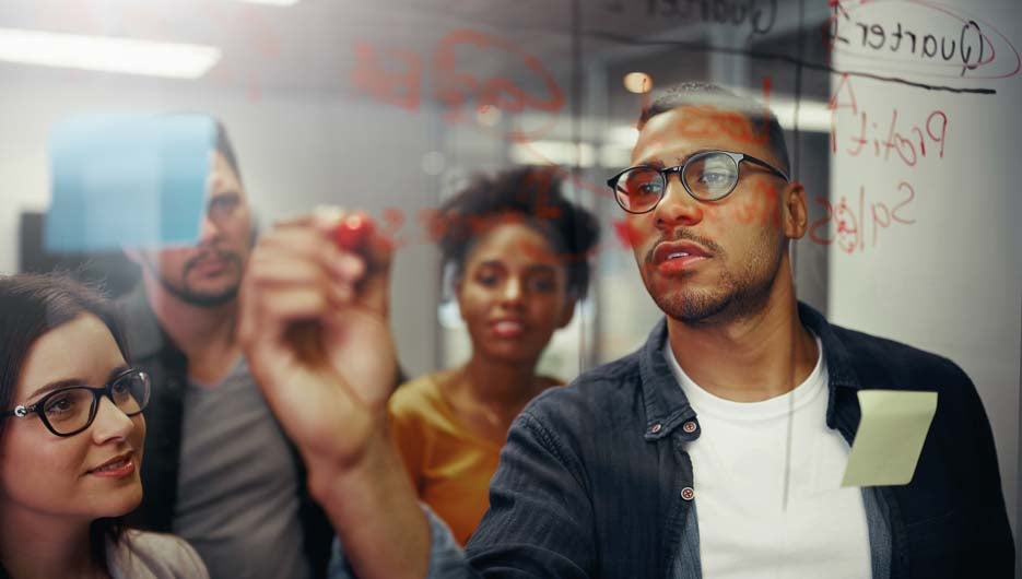Creative team looking at young businessmen writing on glass board in office